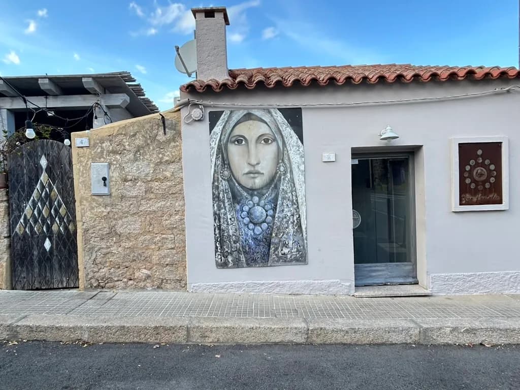 Terracotta-roofed house in San Pantaleo with a painted portrait of a veiled Sardinian woman on its whitewashed wall, beside a wrought-iron gate and shop doorway.