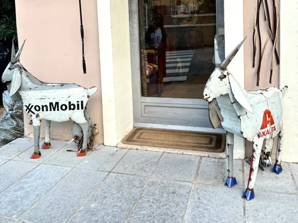 Two recycled metal goat sculptures, made from oil drums with branded logos, stand outside a boutique shopfront in San Pantaleo.
