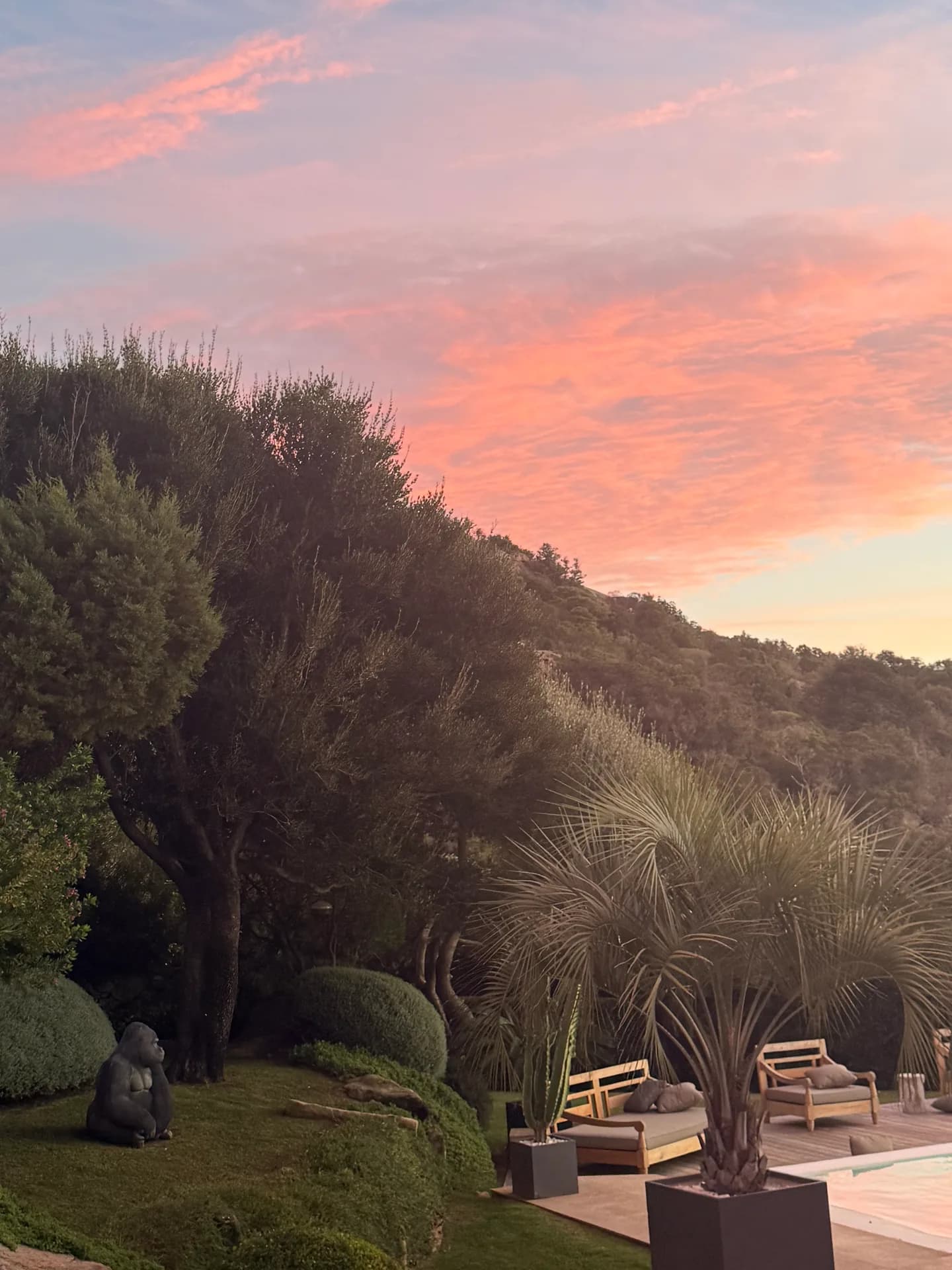 Sunset over a luxury villa garden near Costa Smeralda, with a pink sky above wooded hills, palm plants, outdoor sofas, a pool edge and a gorilla statue on the lawn.