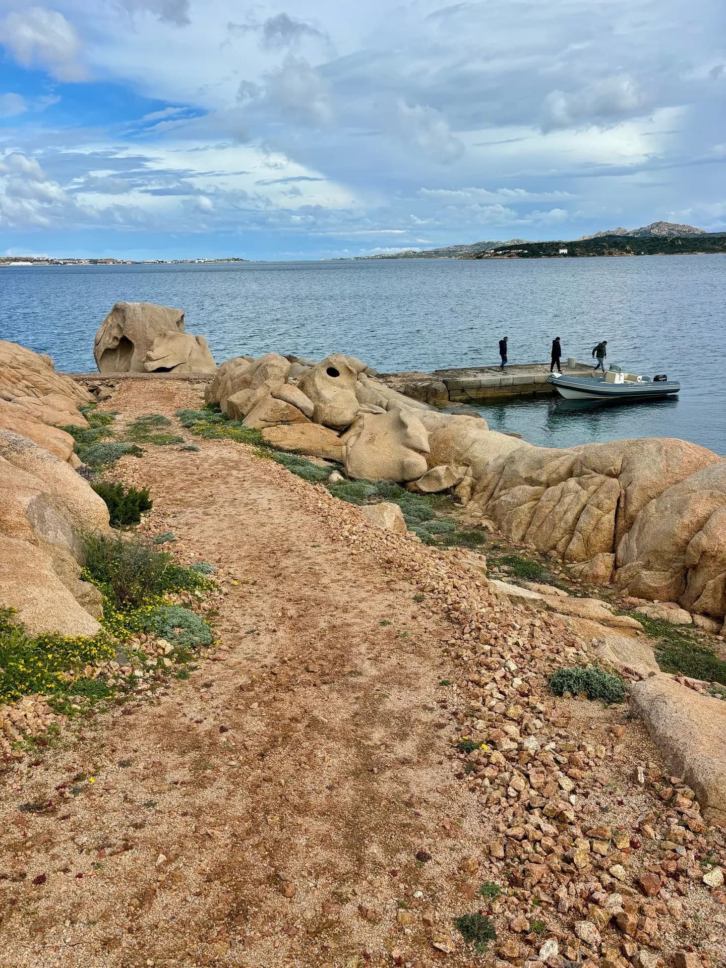 A gravel path winds through wind-eroded granite boulders towards a small jetty on Sardinia's Costa Smeralda coastline, where people board a moored motorboat, with distant hills across the bay.