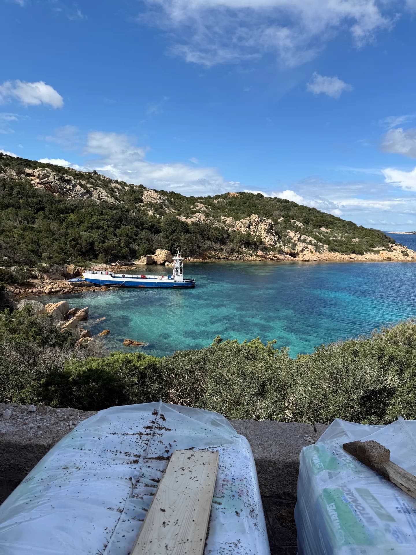 Turquoise cove on Sardinia's coast with a small boat anchored near rocky, scrub-covered hills, building materials covered in plastic sheeting in the foreground.