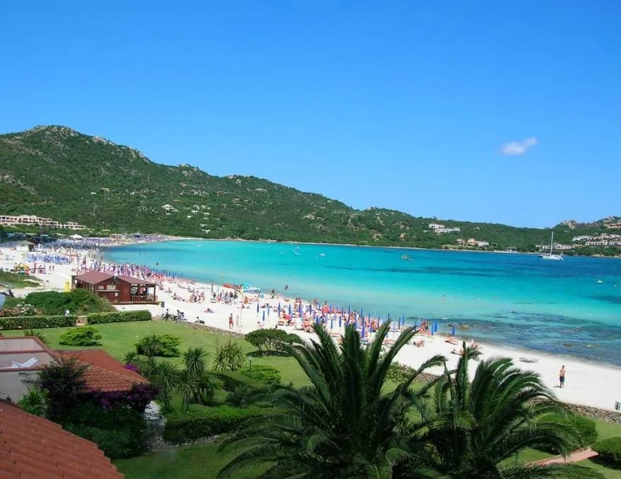 Crowded beach on Sardinia's Costa Smeralda with rows of sun umbrellas along a turquoise bay, green hills and villas in the background.