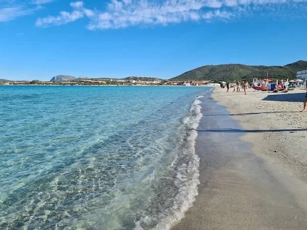 Turquoise sea lapping a sandy Sardinian beach, with hills, a distant coastal town and a few sunbathers under a clear blue sky.