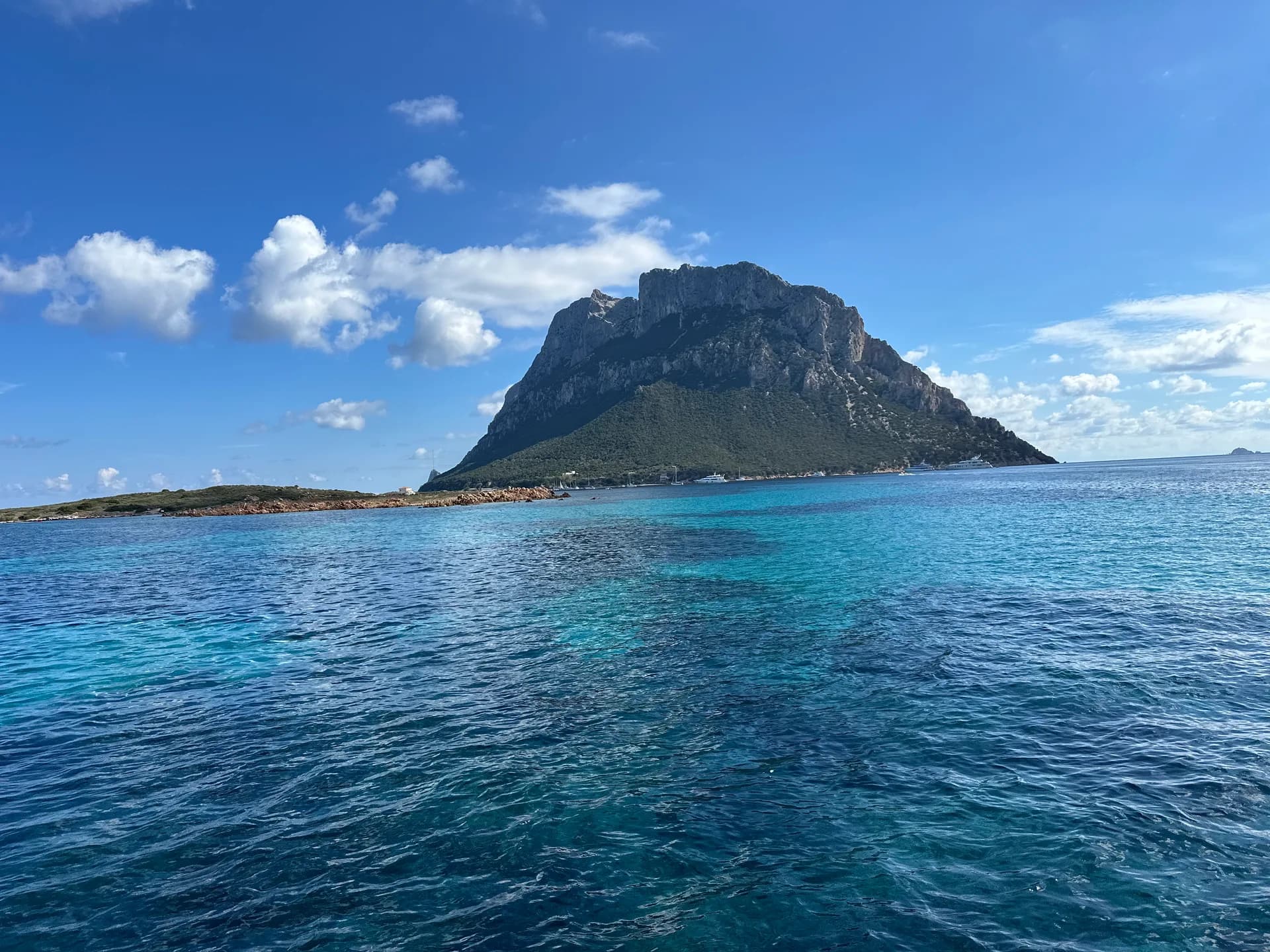 Turquoise sea with the rocky limestone peak of Tavolara island rising above the coast near Sardinia's Costa Smeralda, under a blue sky with scattered clouds and boats moored near the shore.