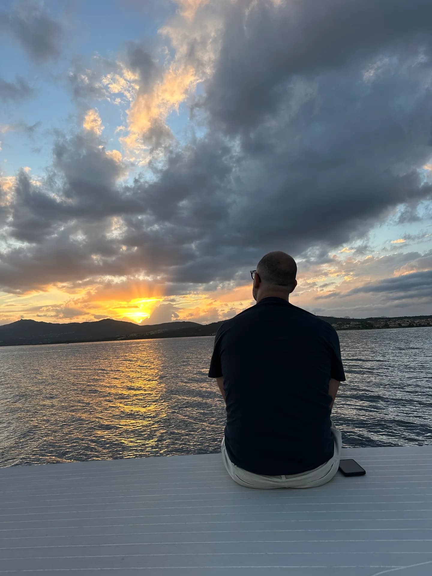 A man sits quietly on a boat deck, watching the sun set over the sea and distant hills near Costa Smeralda, Sardinia, beneath a dramatic cloudy sky.