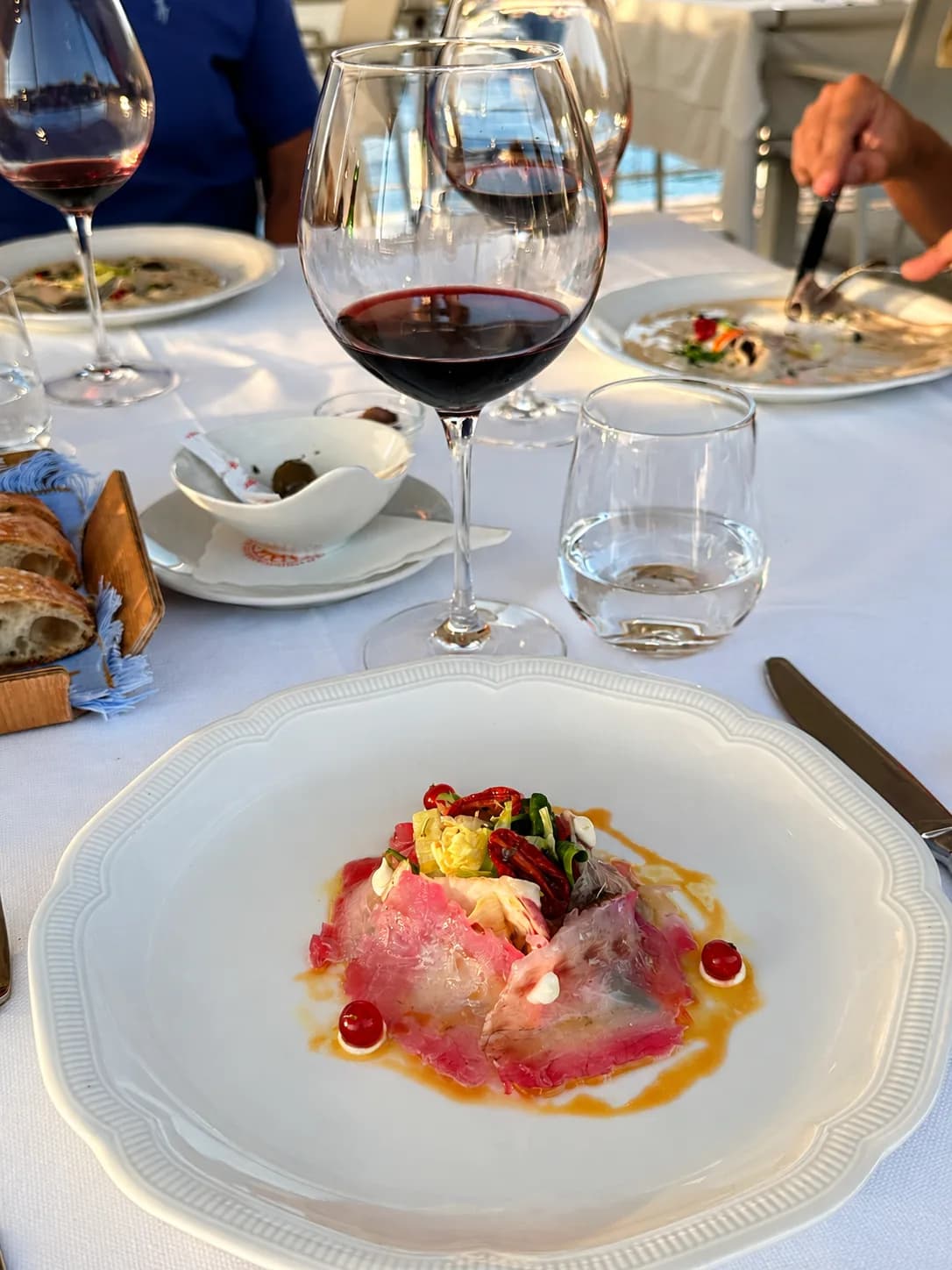 Plated starter of thinly sliced raw fish with leaves, dried tomato and red berries on a white plate, beside a glass of red wine, water and bread on a white-clothed table