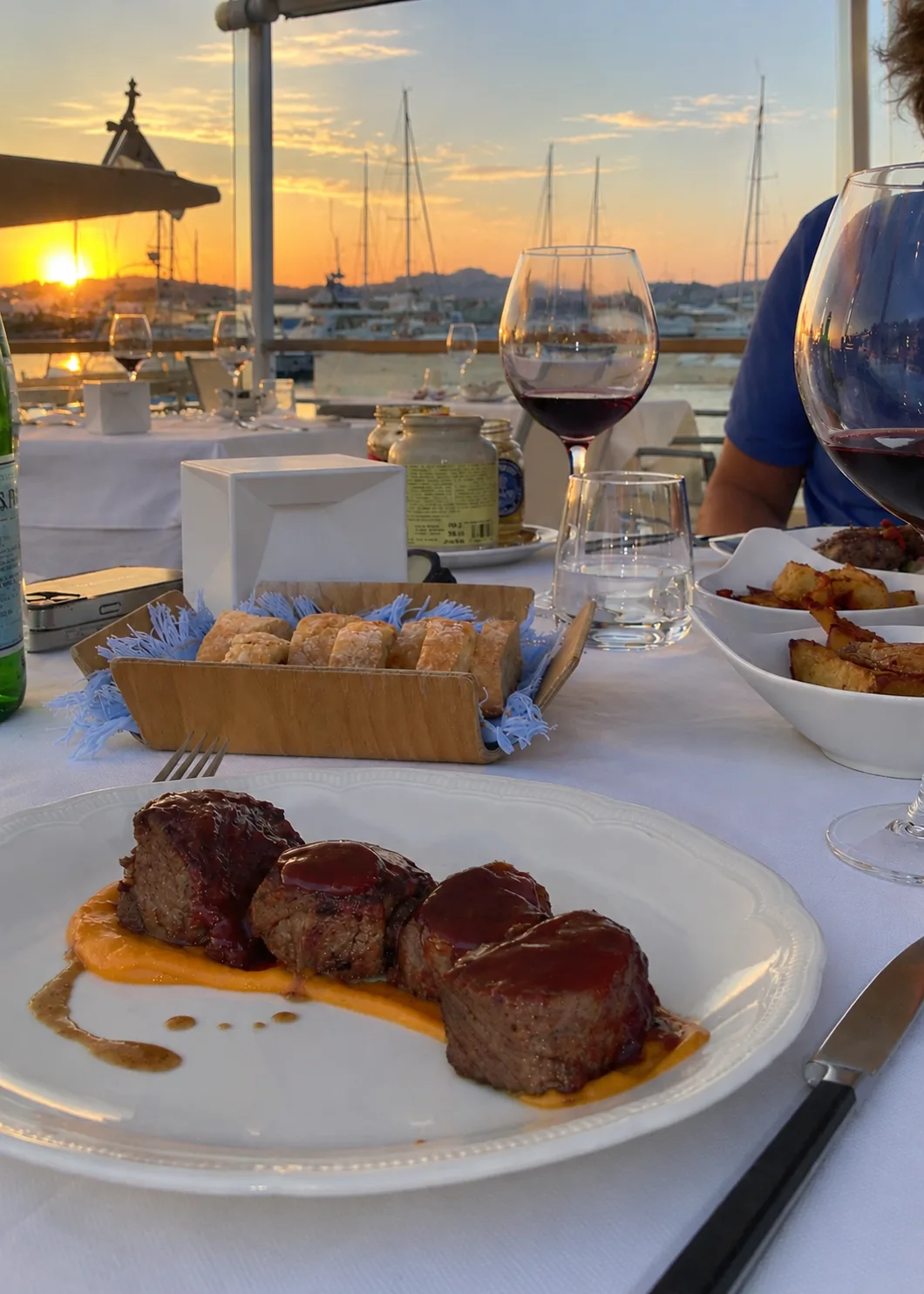 Glazed medallions of beef on a puréed orange sauce, with red wine, bread and roast potatoes on a white-clothed table overlooking a marina with yacht masts at sunset