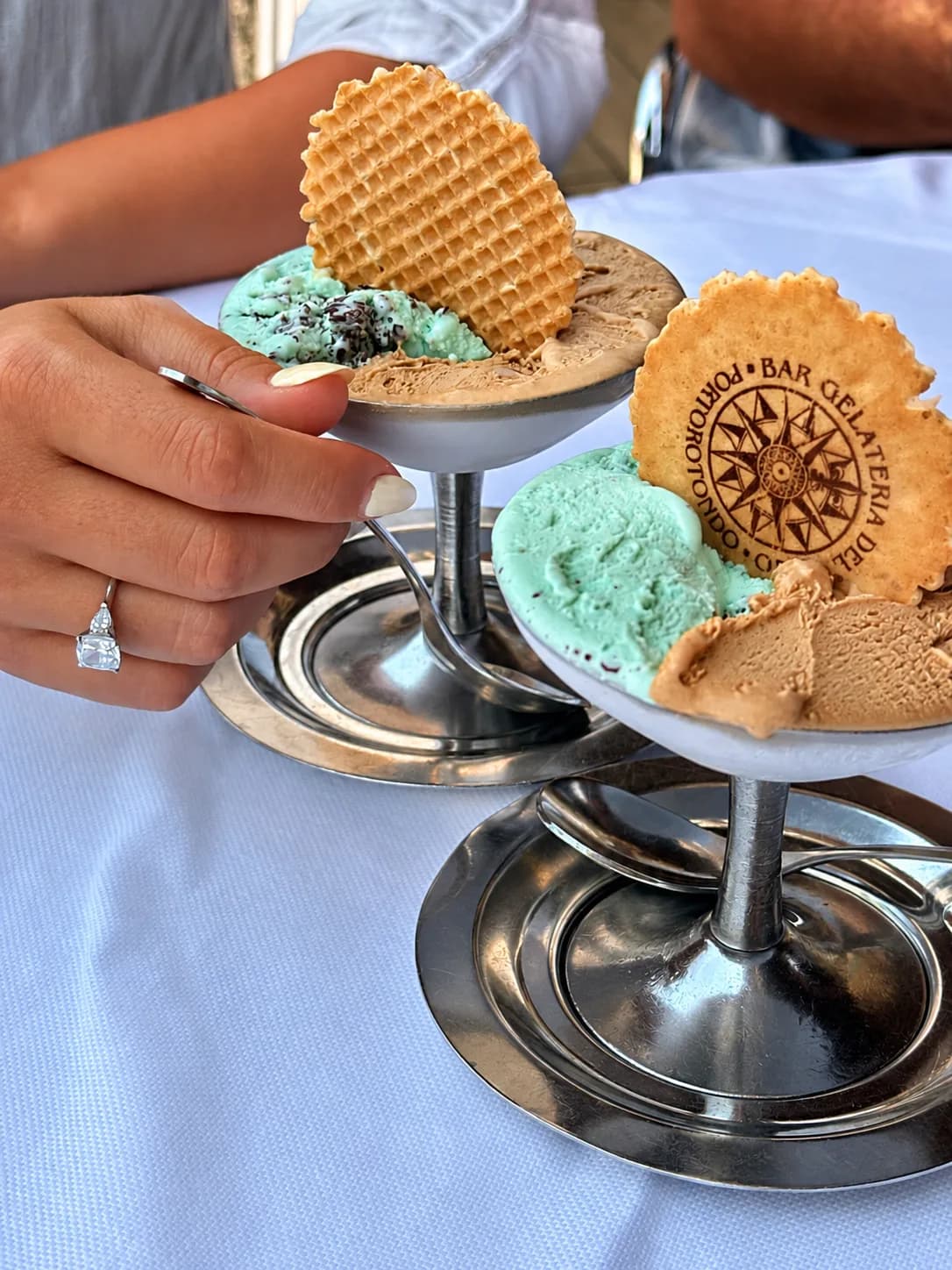 Two metal pedestal dishes of mint-chocolate-chip and coffee-coloured ice cream with wafers, one stamped with a compass design and Porto Rotondo text; a hand holds a spoon.