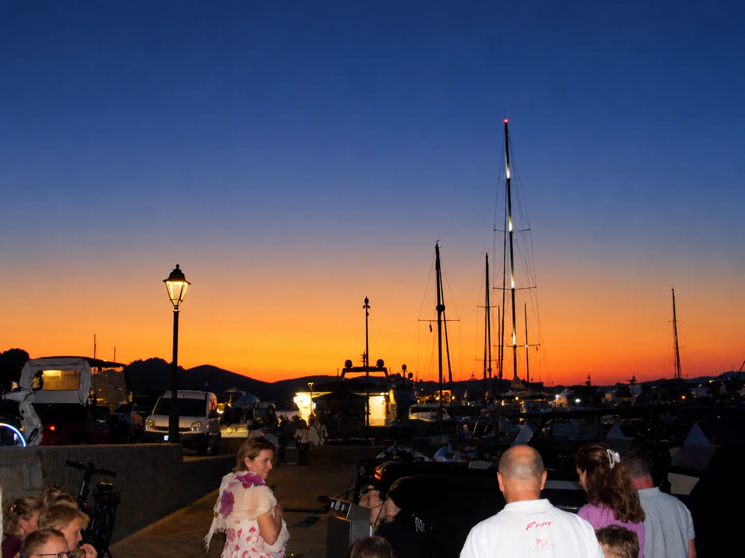 Diners and passers-by on a harbour quayside at dusk, with yacht masts silhouetted against an orange and deep blue sky and a lit street lamp beside the water