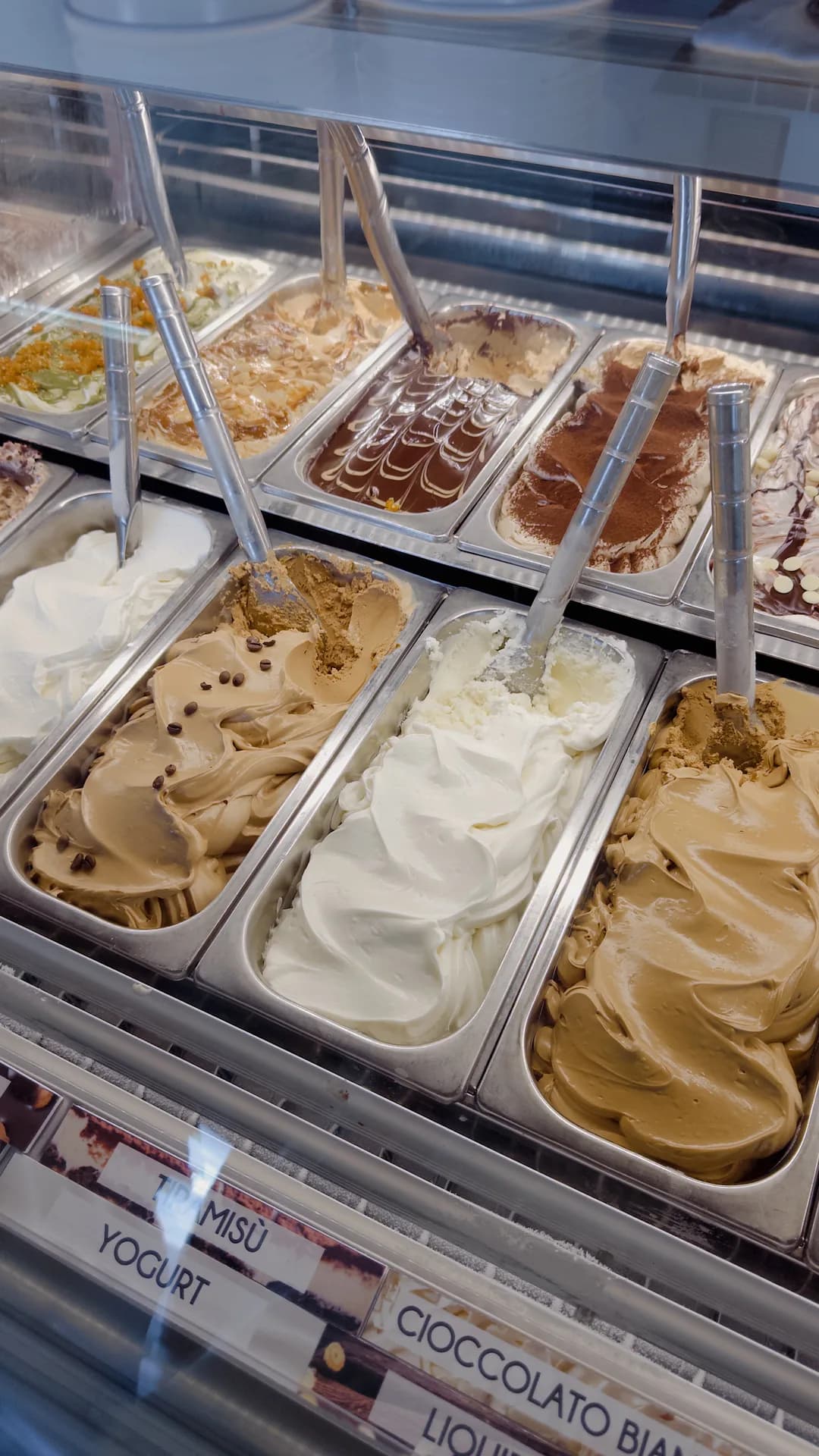 Glass-fronted gelato display with metal tubs of swirled coffee, white cream, chocolate and cocoa-dusted flavours, each with a scoop, and labels reading Tiramisù, Yogurt and Cioccolato bianco.