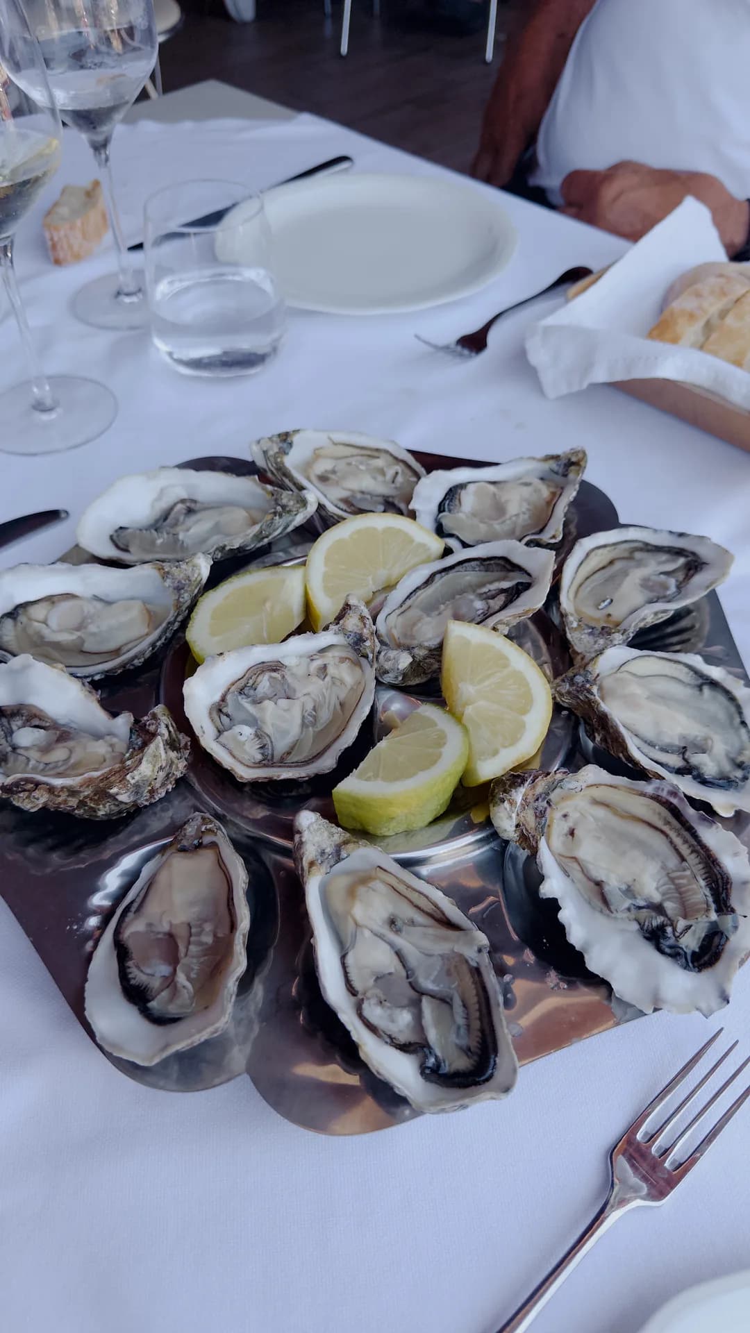 Platter of a dozen fresh oysters on the half shell with lemon wedges, set on a white-clothclad table with wine glasses, a bread basket and cutlery.