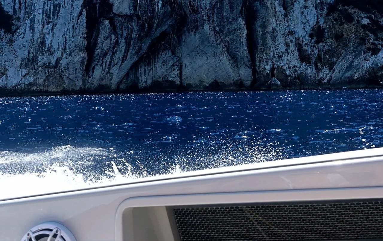 View from a boat skimming deep blue water beneath towering limestone cliffs along the Sardinian coast.
