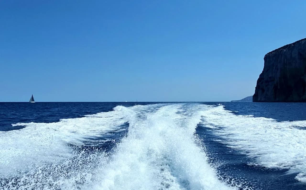 Frothy white wake trailing a boat across deep blue Sardinian sea, with a distant sailboat and a rocky headland on the horizon.