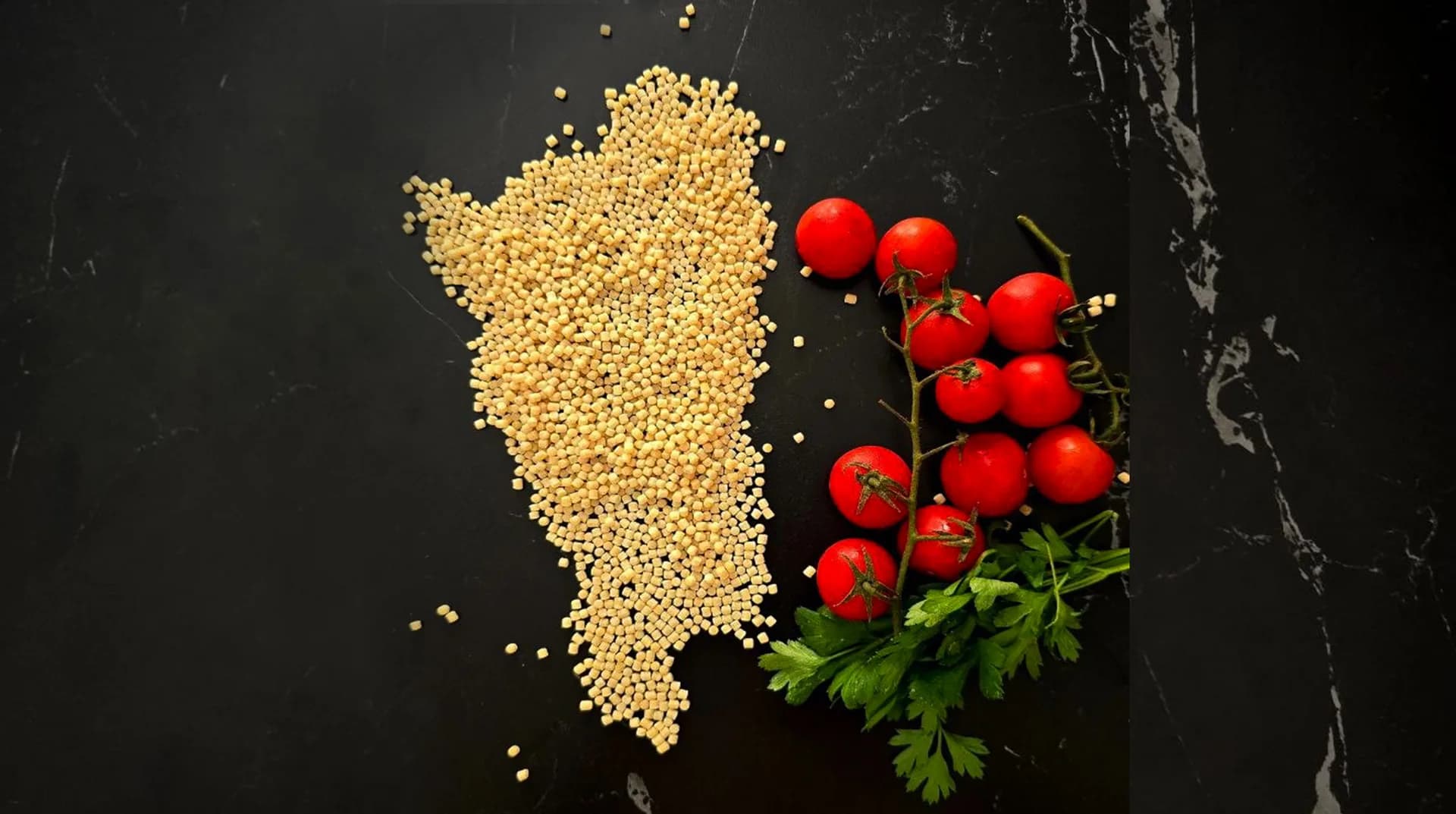 Dried fregola pasta grains arranged in the shape of Sardinia on a dark surface, beside vine tomatoes and fresh parsley