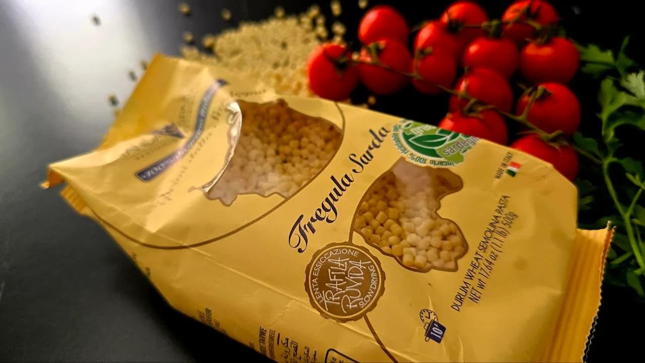A yellow packet of Fregula Sarda pasta beside loose grains, cherry tomatoes and parsley on a dark surface