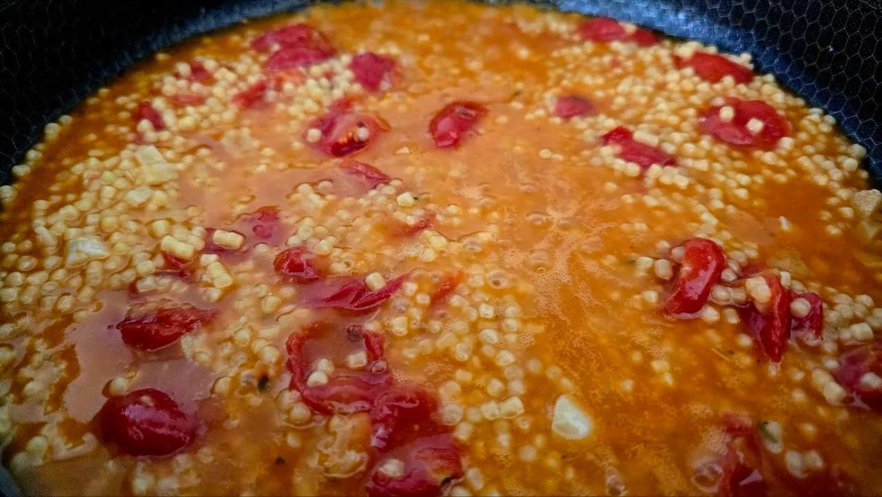 Close-up of fregola sarda simmering in a tomato broth with chunks of cherry tomatoes and garlic in a non-stick pan