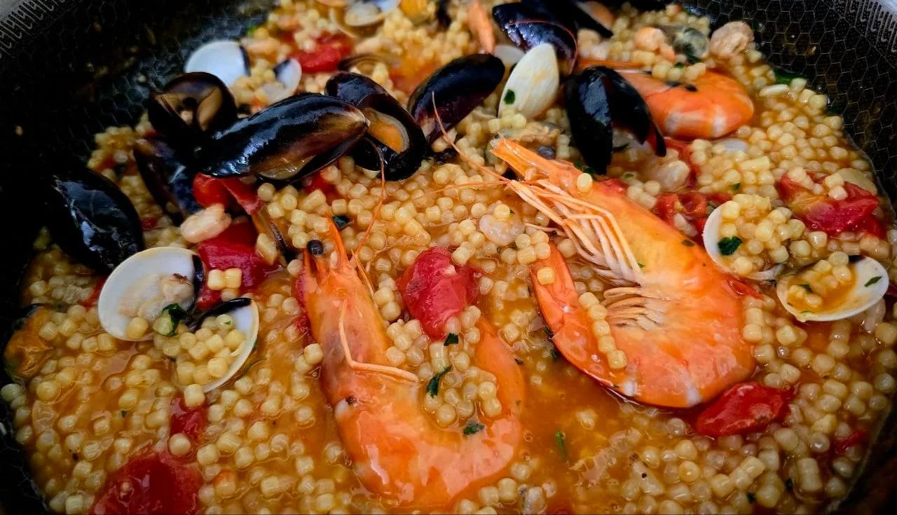 Close-up of fregola sarda with prawns, mussels and clams simmered in a tomato broth, in a pan.