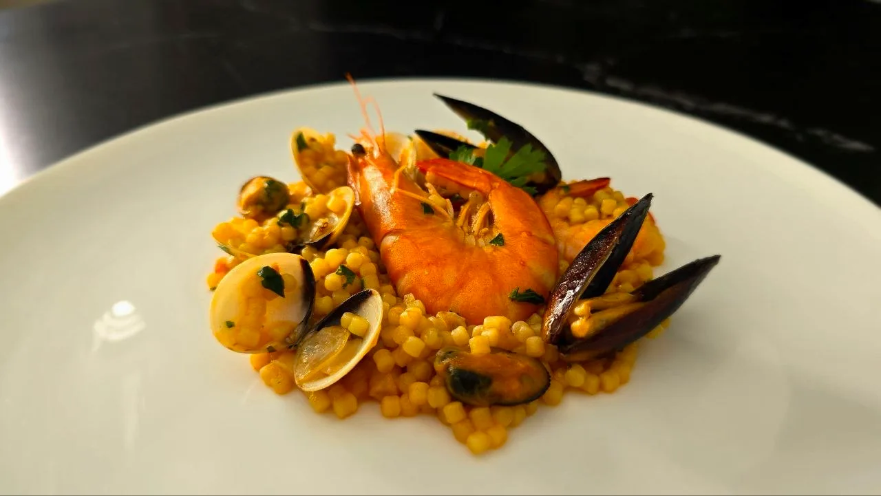 A plate of saffron-coloured fregola sarda topped with a whole prawn, clams, mussels and chopped parsley