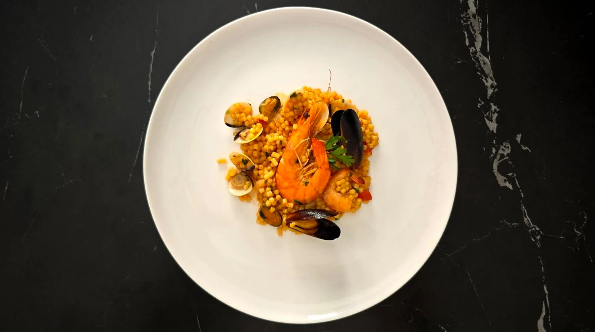 Overhead view of a white plate holding fregola sarda pasta topped with prawns, mussels and clams in a tomato-tinted broth, set on a dark marble surface.