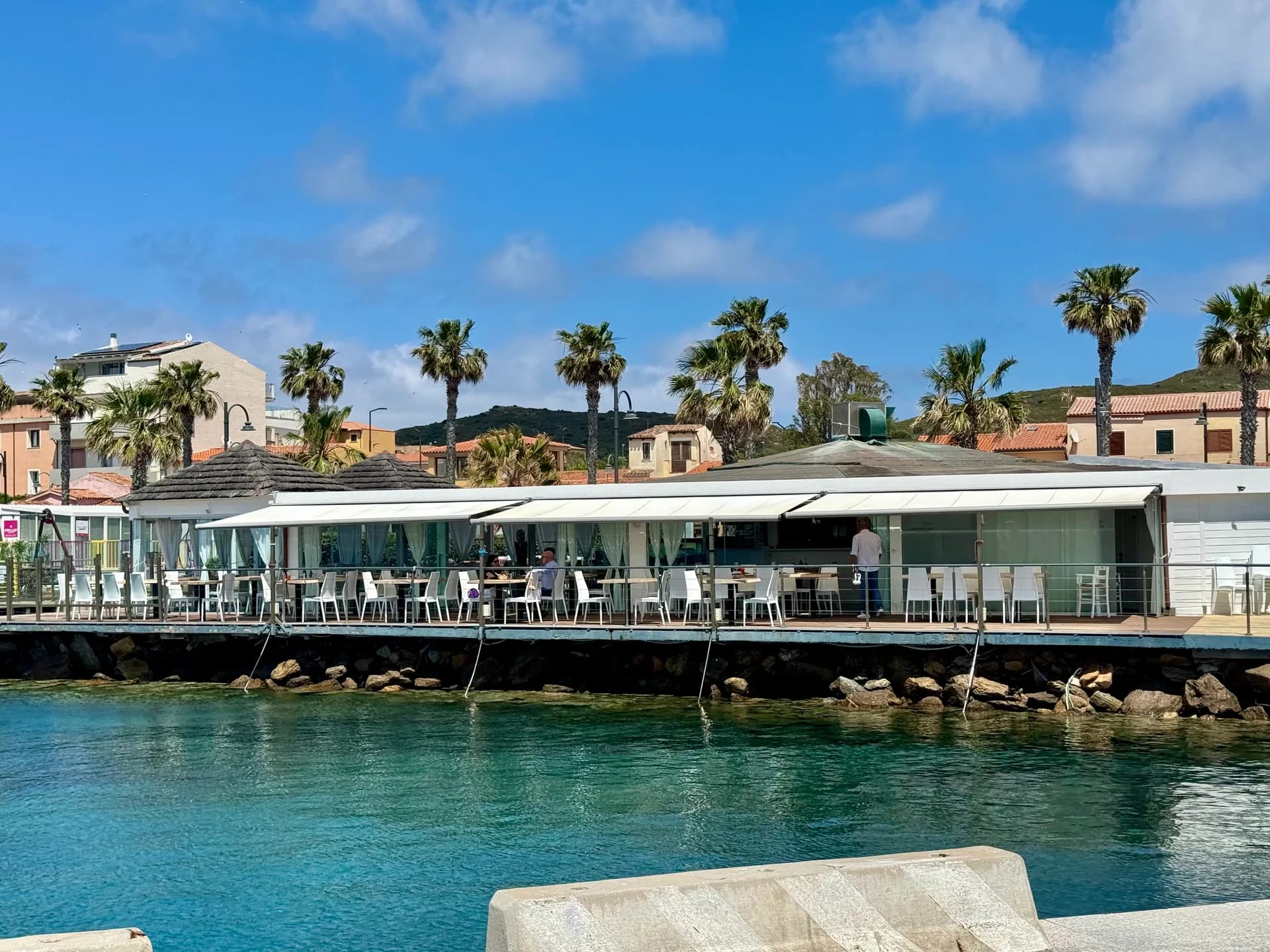 Waterfront restaurant terrace with white chairs and awnings above turquoise water, with palm trees, terracotta-roofed buildings and green hills under a blue sky