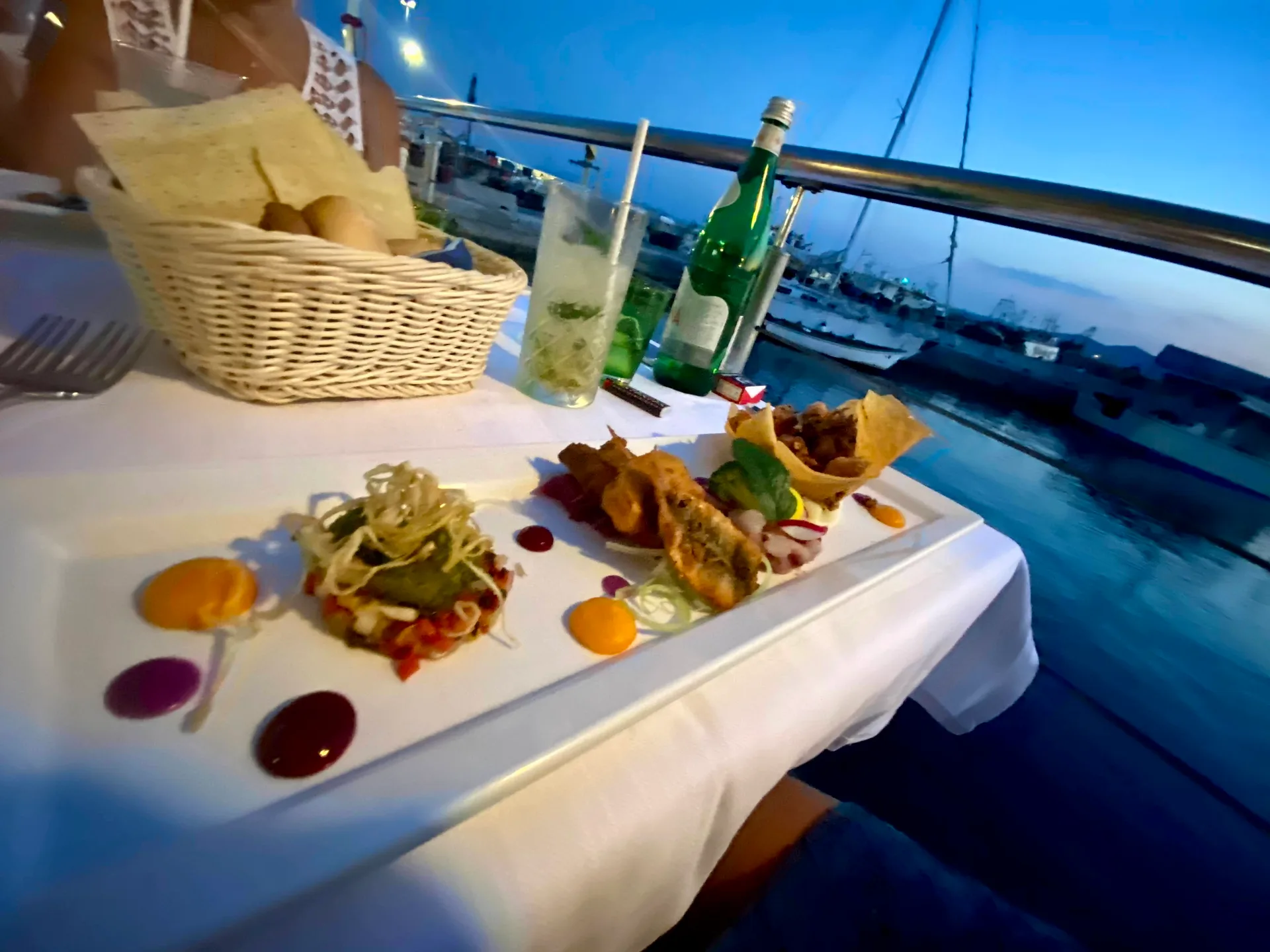 Plated seafood starter with fried fish, a crisp basket and colourful sauces on a white tablecloth, beside a bread basket, a mojito and a bottle, with boats in a harbour at dusk.