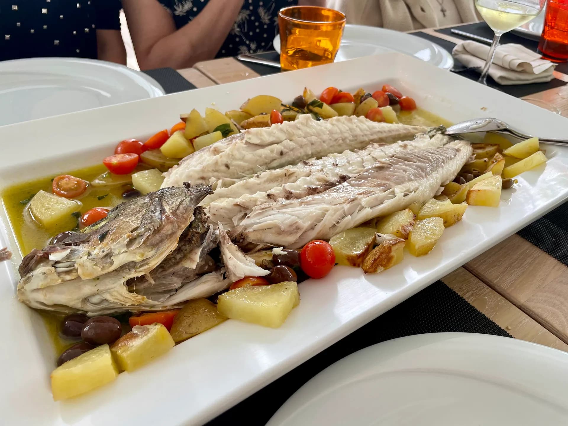 Whole cooked fish, filleted and served on a long white platter with potato chunks, halved cherry tomatoes, olives and herb broth, on a restaurant table with orange glasses and white wine.