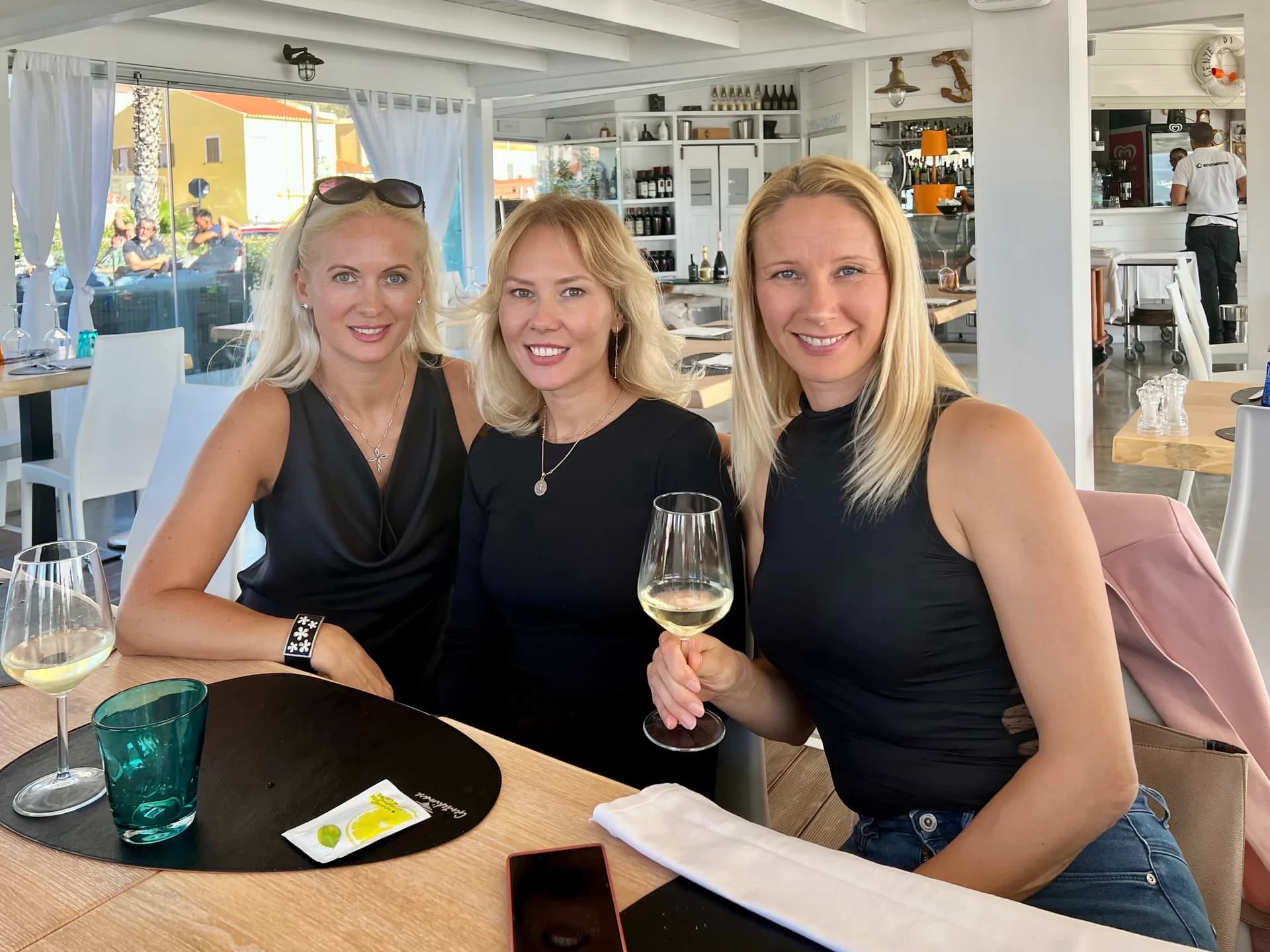 Three smiling blonde women in black tops sit at a wooden table with glasses of white wine in a bright, white nautical-style restaurant, with a seafront view behind.