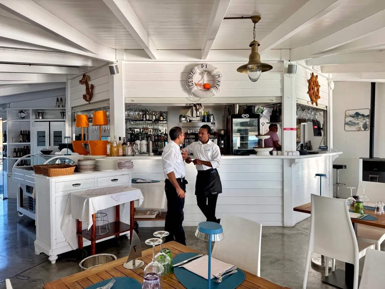 Two waiters in white shirts and black aprons talk in front of a white wooden bar with a lifebuoy reading 'Gente di Mare', a brass lamp and set tables in the foreground.