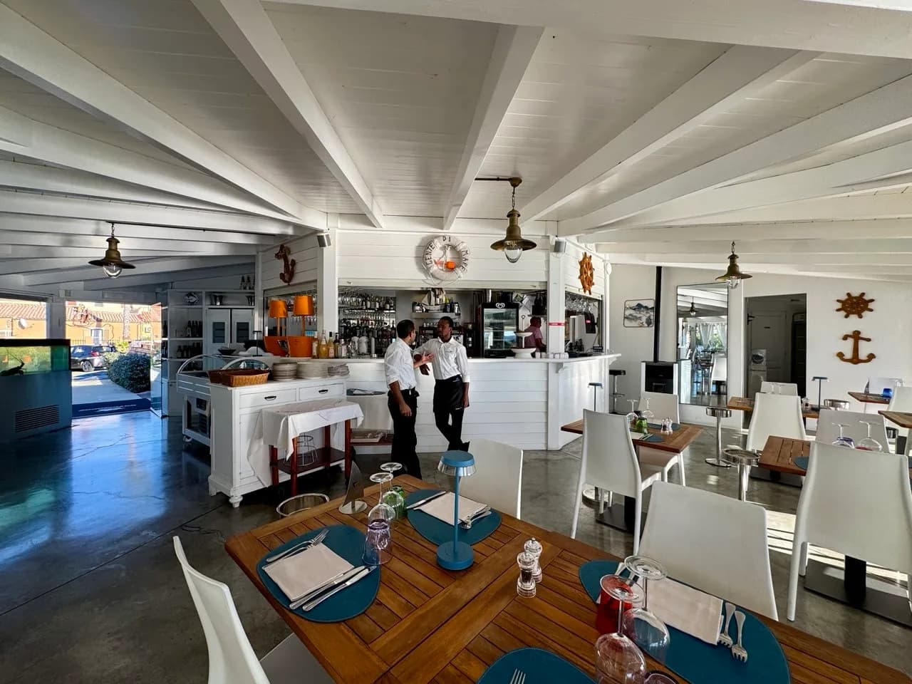 Bright restaurant interior with white-beamed ceiling, nautical decor and a central bar, where two waiters in white shirts talk beside wooden tables laid with blue placemats.