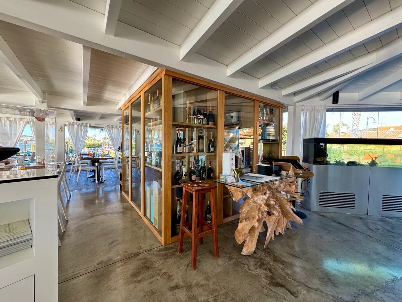 Airy seafront restaurant interior with white beamed ceiling, a glass wine cabinet, a driftwood-based reception desk, a lobster tank and tables by sea-view windows