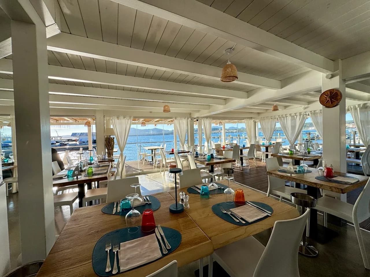 Bright waterfront restaurant interior with white beamed ceiling, wooden tables set with blue placemats and coloured glasses, and white curtains framing sea and hills beyond.