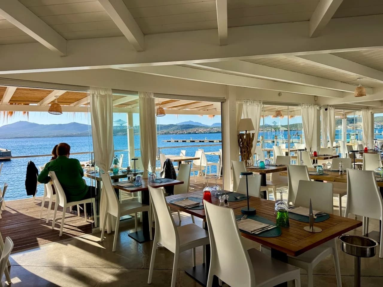 Bright waterfront restaurant with white chairs and set wooden tables, glass doors opening onto a terrace, a diner looking out over blue sea towards distant hills.
