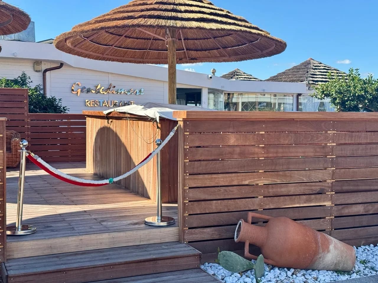 Wooden deck entrance to a white seafront restaurant with a red rope barrier, thatched parasols, a terracotta amphora on white pebbles and a prickly pear cactus under blue sky