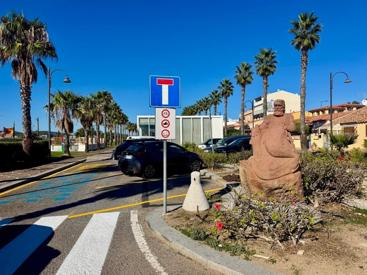 Seafront road lined with palm trees under a clear blue sky, with a dead-end sign, parked cars and a carved reddish stone statue of a bearded man in a flowerbed.