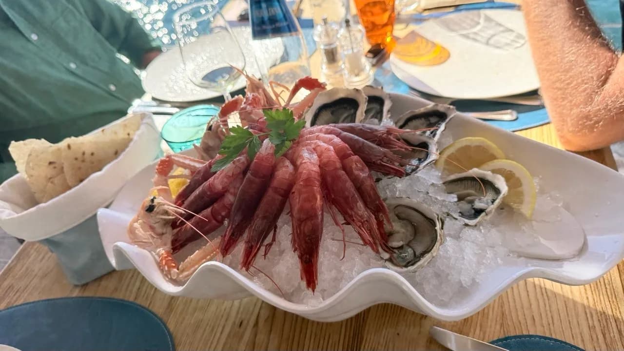 Shell-shaped white dish of raw red prawns, langoustines and oysters on crushed ice with lemon slices and parsley, on a sunlit restaurant table beside a basket of flatbread.