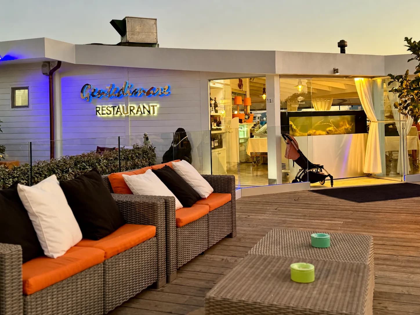 Dusk view of a white seafront restaurant with illuminated 'Gente di mare Restaurant' sign, glass frontage, a fish tank inside and a wooden deck with orange-cushioned wicker sofas.