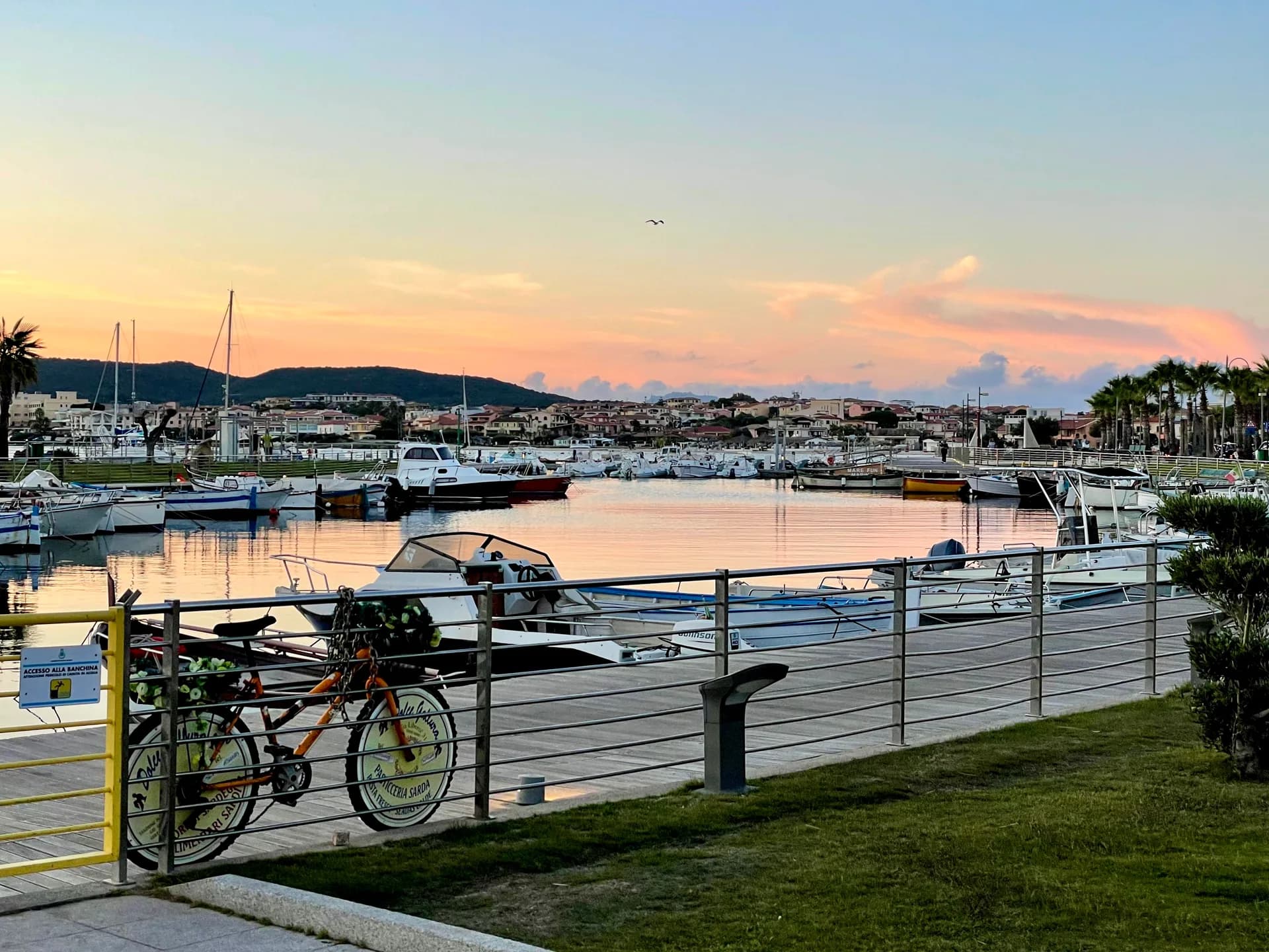 Sunset over Golfo Aranci marina, boats moored along the harbour, a bicycle chained to railings on the promenade, with hills and town beyond.