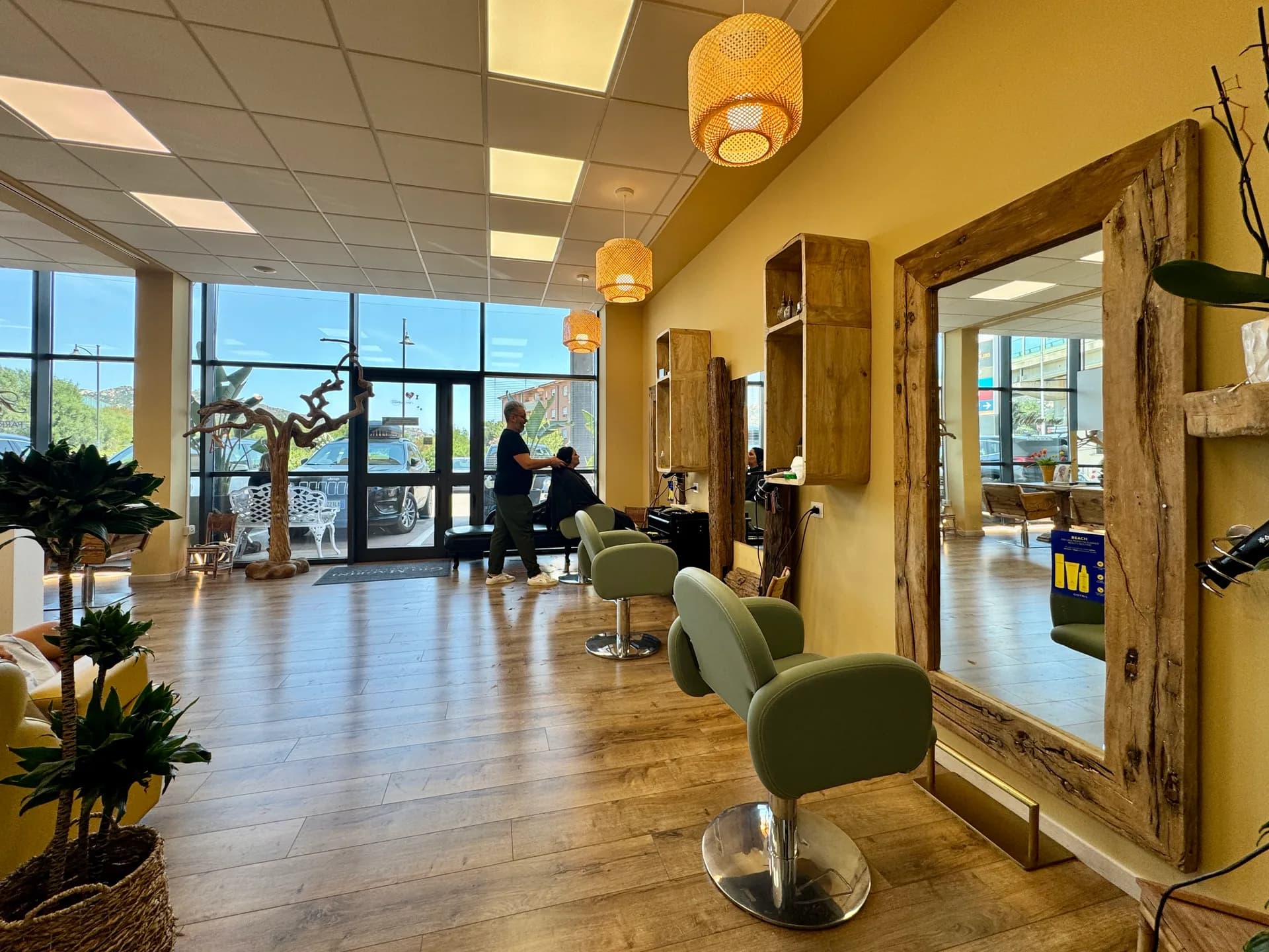 Modern hairdressing salon with green swivel chairs, driftwood-framed mirrors and woven pendant lights; a stylist attends a client by the glass entrance.