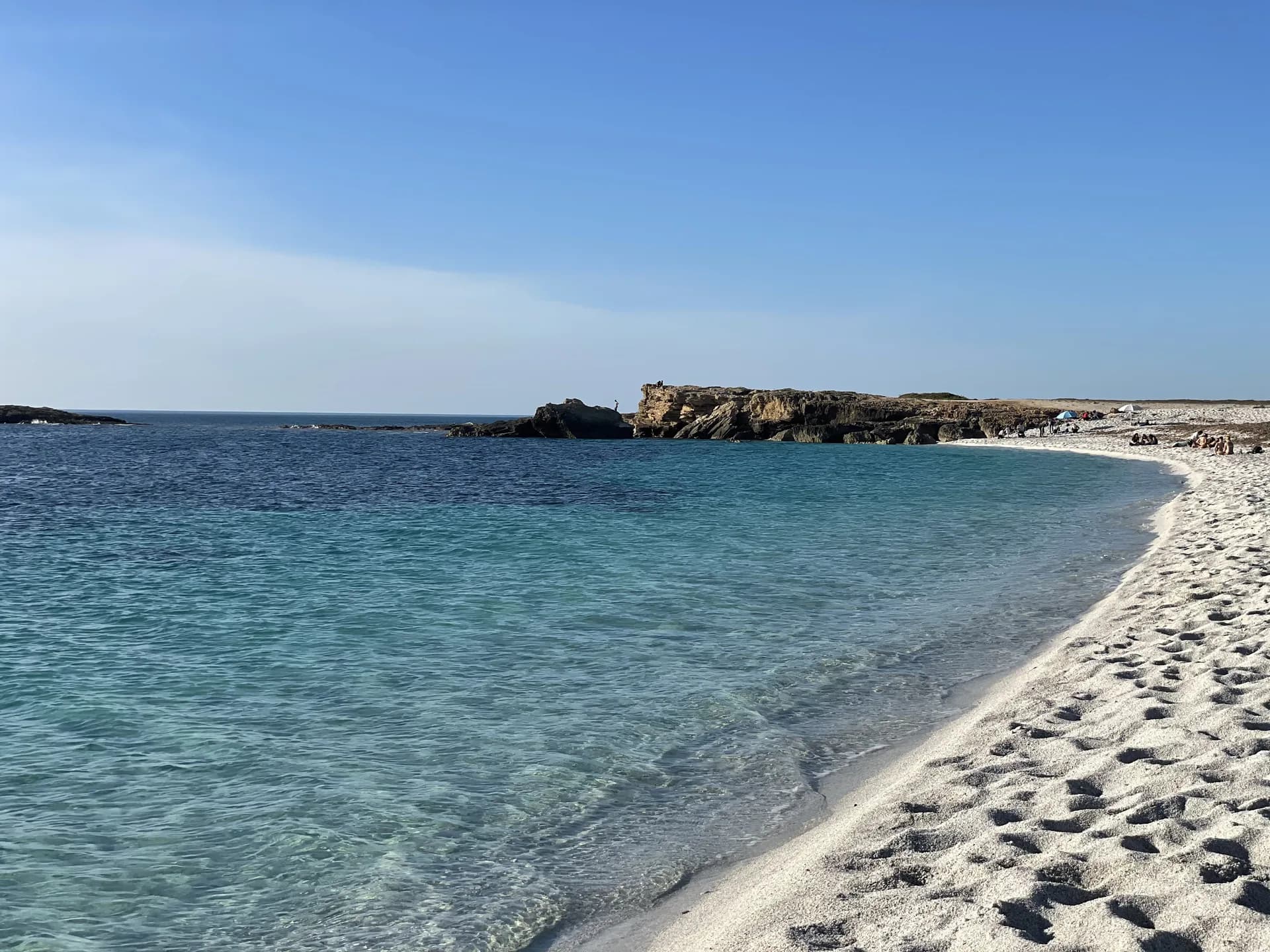 Clear turquoise water curving along the white quartz-grain shore of Is Arutas, with a low rocky headland beyond