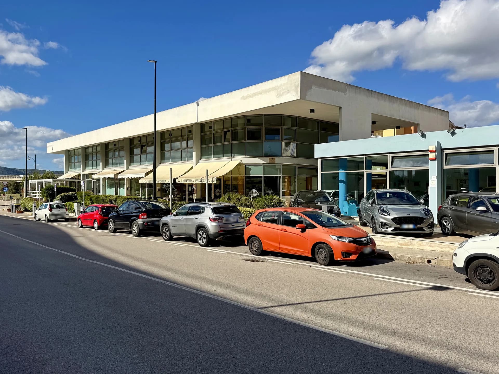 Zweistöckiges weißes modernes Gebäude mit cremefarbenen Markisen mit der Aufschrift „Caffetteria Pasticceria“, davor an der Straße geparkte Autos und rechts ein Ausstellungsraum mit blauer Front unter strahlend blauem Himmel