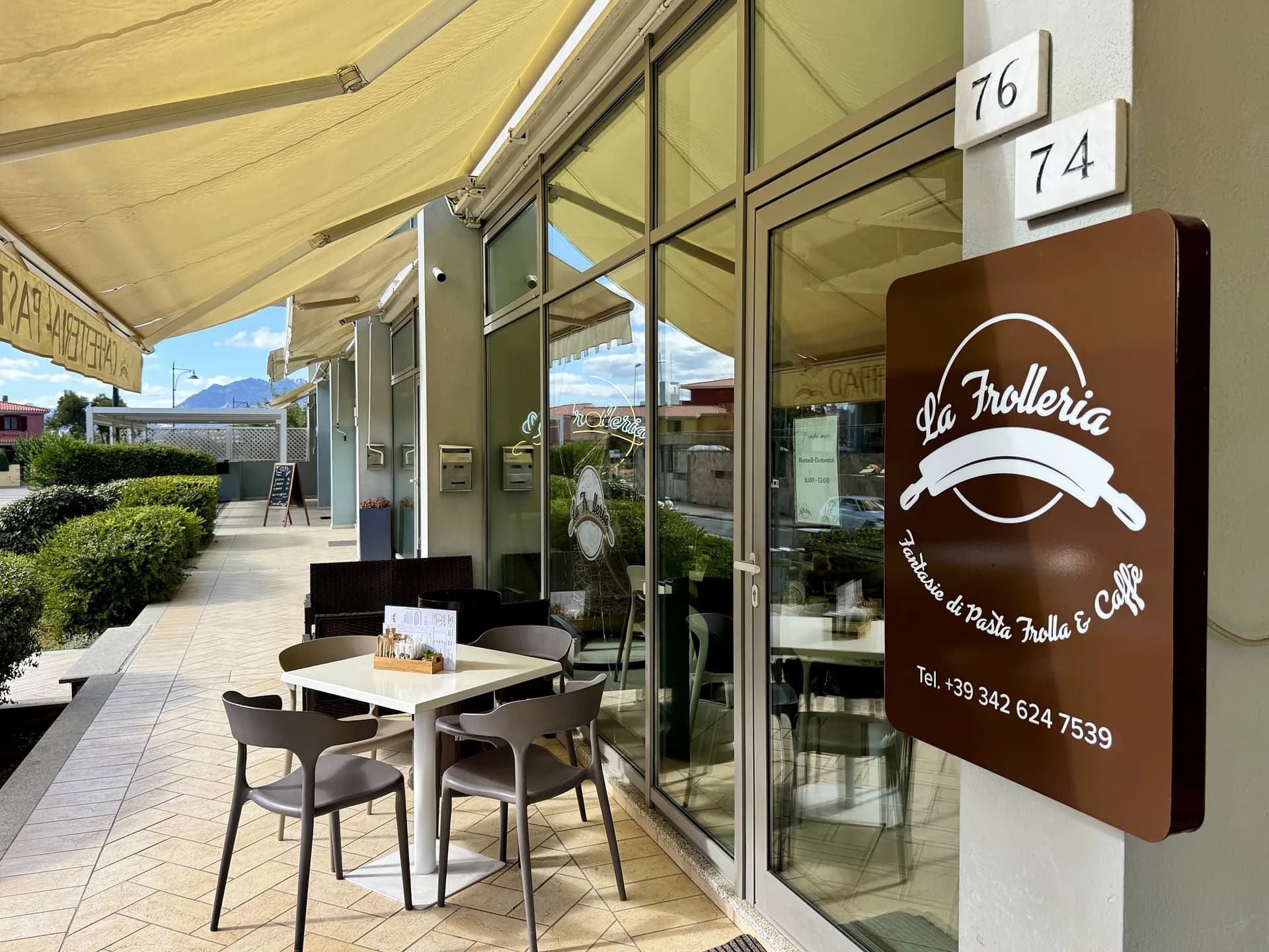 Café-Fassade mit braunem Schild „La Frolleria“ mit einem Nudelholz, Glastüren, gelber Markise und einem kleinen Tisch mit grauen Stühlen auf einem gefliesten Gehweg, dahinter Berge.