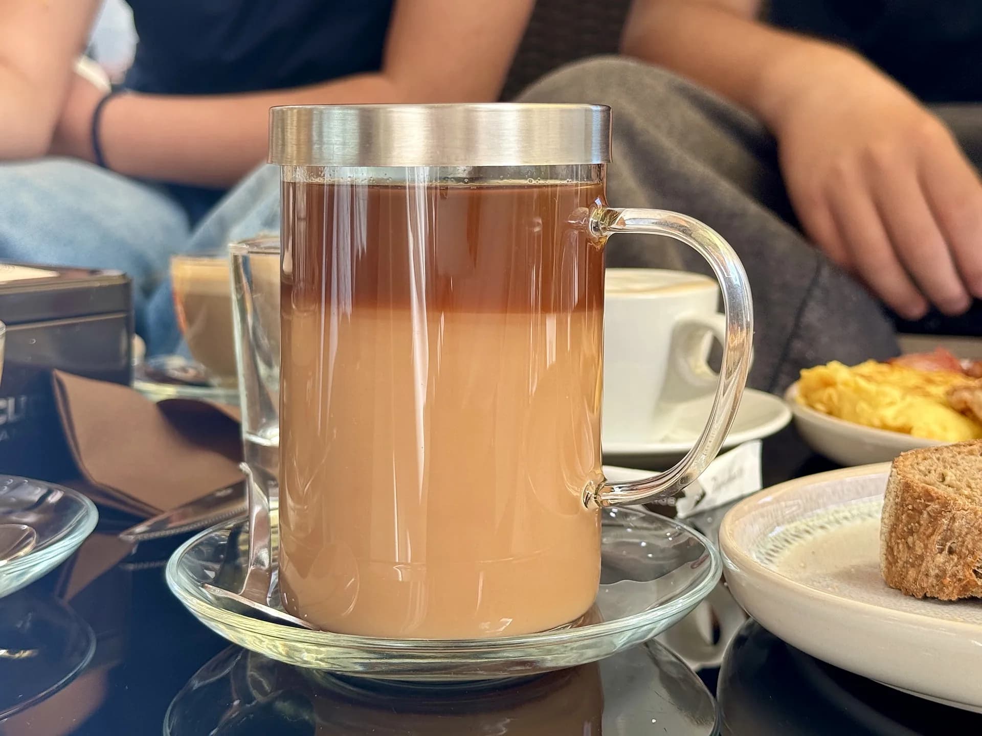 Geschichteter Milchkaffee in einem hohen Glasbecher mit Metalldeckel auf einer Glasuntertasse, dahinter auf dem Café-Tisch Rührei, eine Scheibe Vollkornbrot und ein Cappuccino.