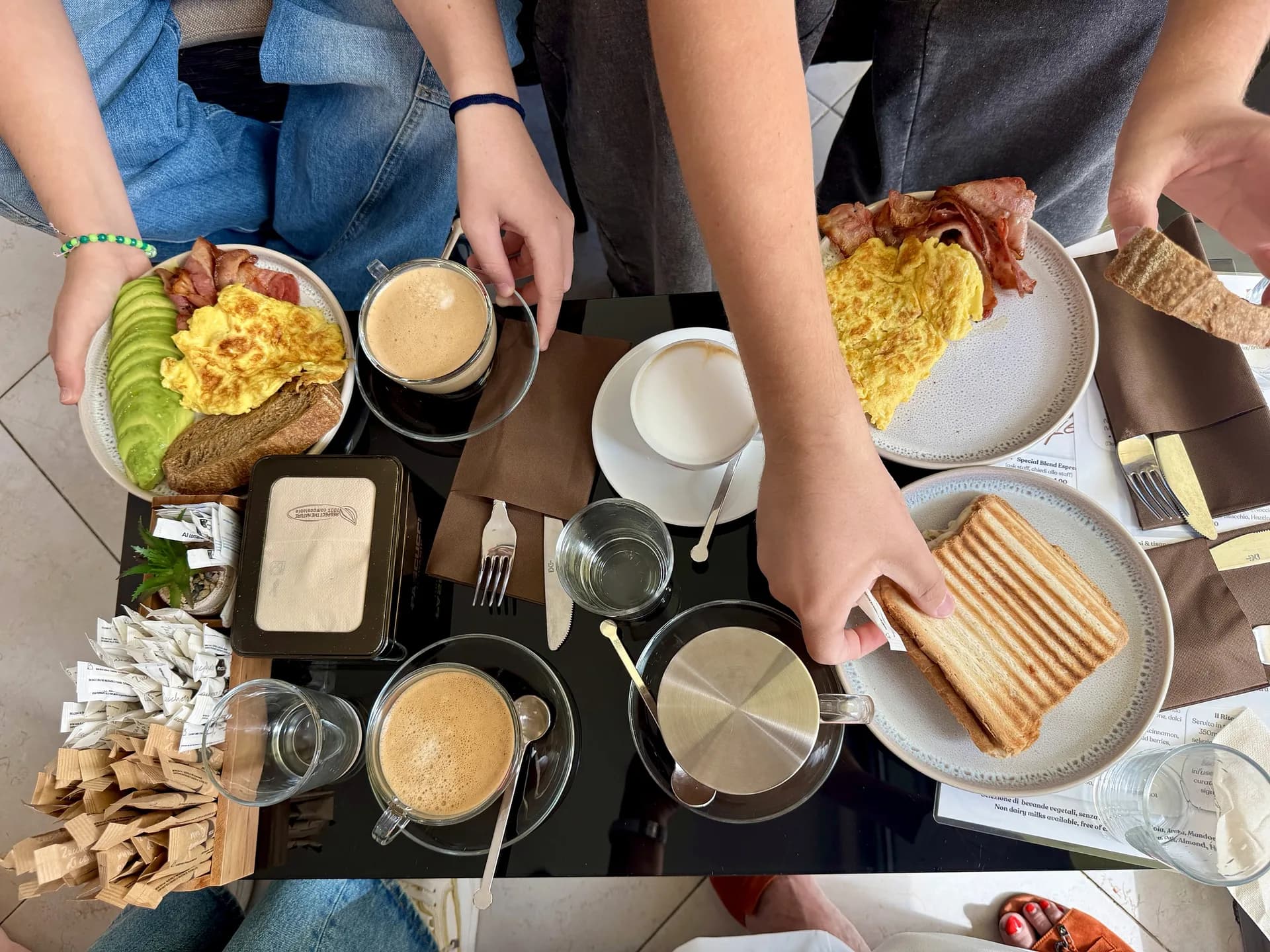 Draufsicht auf einen zum Frühstück gedeckten Café-Tisch: Teller mit Rührei, Speck, Avocado und Toast, ein getoastetes Sandwich, Kaffees, Wassergläser und Zuckertütchen, dazu Hände der Gäste, die zugreifen.