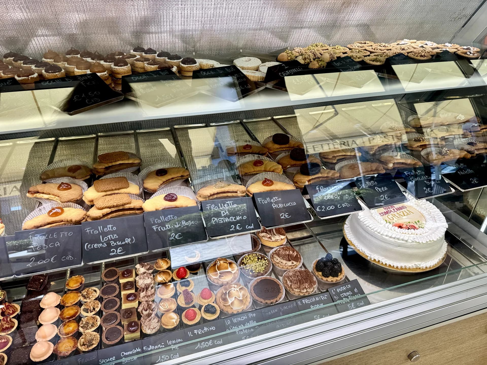 Cremefarbener hängender Korbei-Sessel neben einem Neonschriftzug „La Frolleria“ an einer grünen Heckenwand, durch das Fenster sind Café-Tische, Stühle und eine gelbe Markise zu sehen.