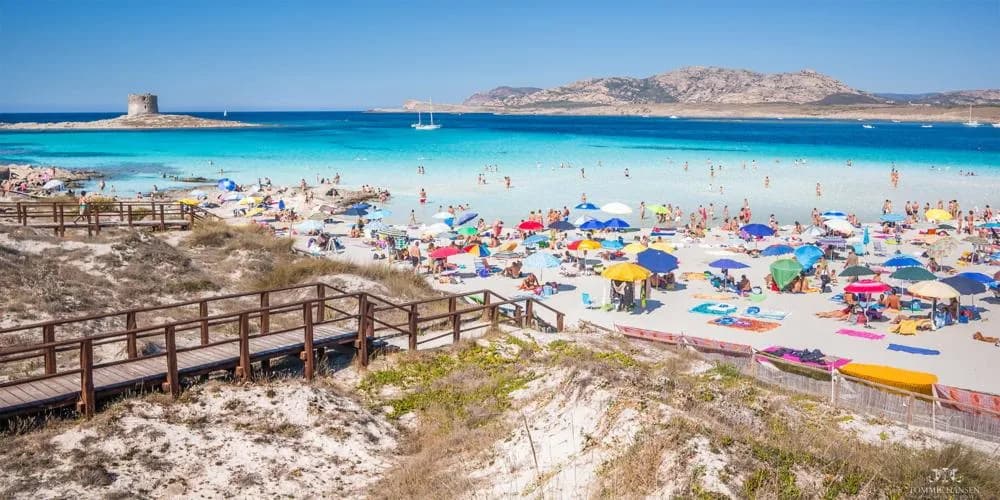 Wooden boardwalk leading down dunes to La Pelosa Beach, Stintino, with crowds, colourful umbrellas, turquoise sea, a Spanish watchtower and Asinara island in the distance.