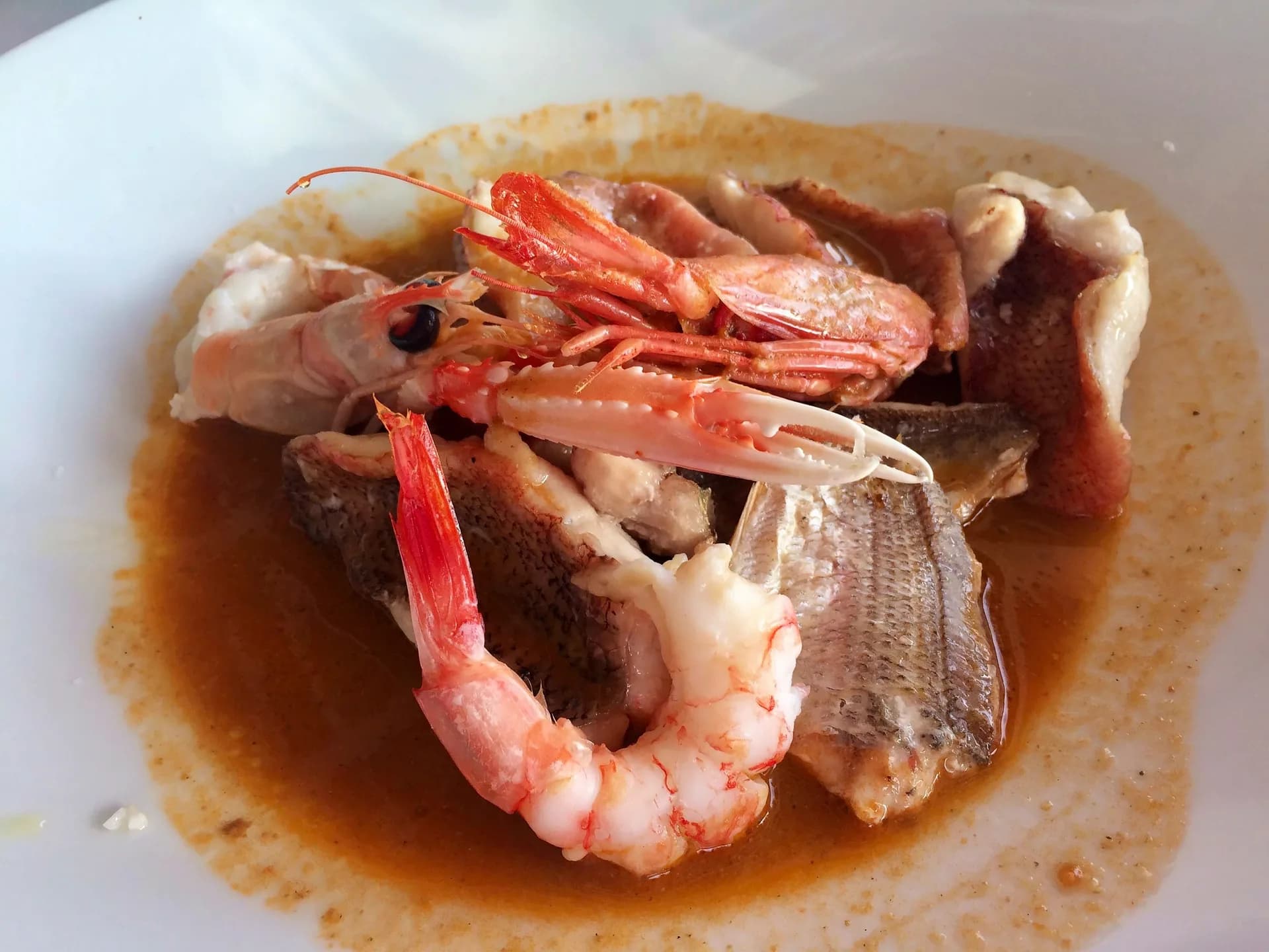 Primo piano di una ciotola bianca con zuppa di pesce con filetti, uno scampo dalla chela chiara e un gambero rosa in un ricco brodo arancio-brunastro