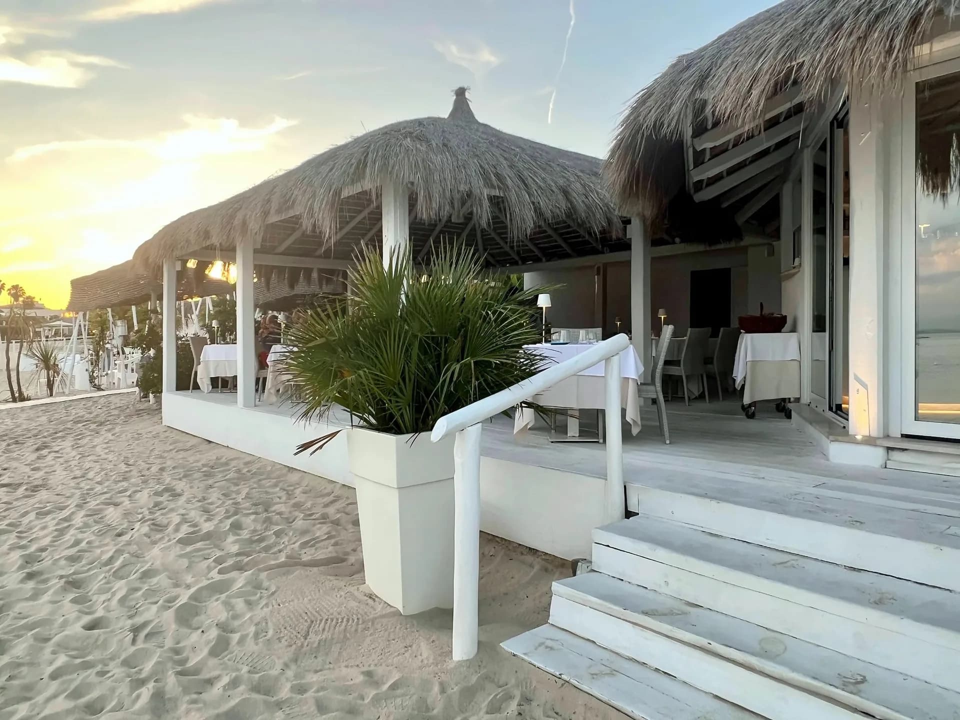 Ristorante in spiaggia con tetto di paglia e tavoli con tovaglie bianche su una pedana di legno chiaro, con gradini bianchi e una palma in vaso, sulla sabbia al tramonto