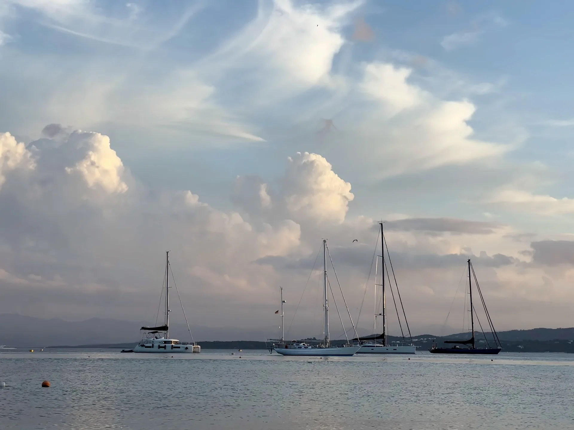 Quattro barche a vela e un catamarano all'ancora su un'acqua calma e argentea al crepuscolo, con imponenti nuvole illuminate di rosa e colline lontane sullo sfondo.