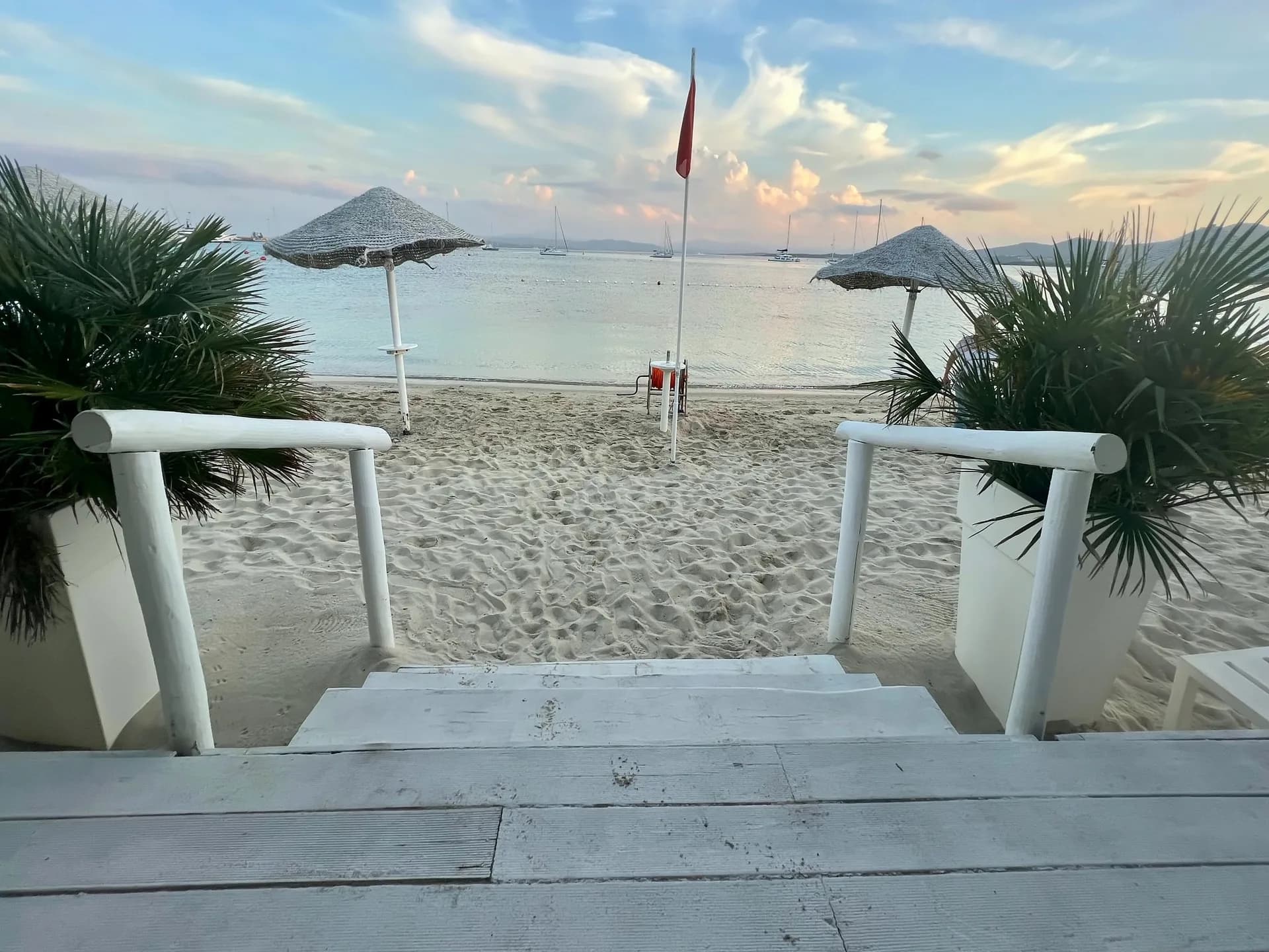 Gradini di legno bianco con corrimano di tronchi e palme in vaso scendono verso una spiaggia sabbiosa con ombrelloni di paglia, una bandiera rossa e barche a vela ormeggiate sull'acqua calma al crepuscolo.