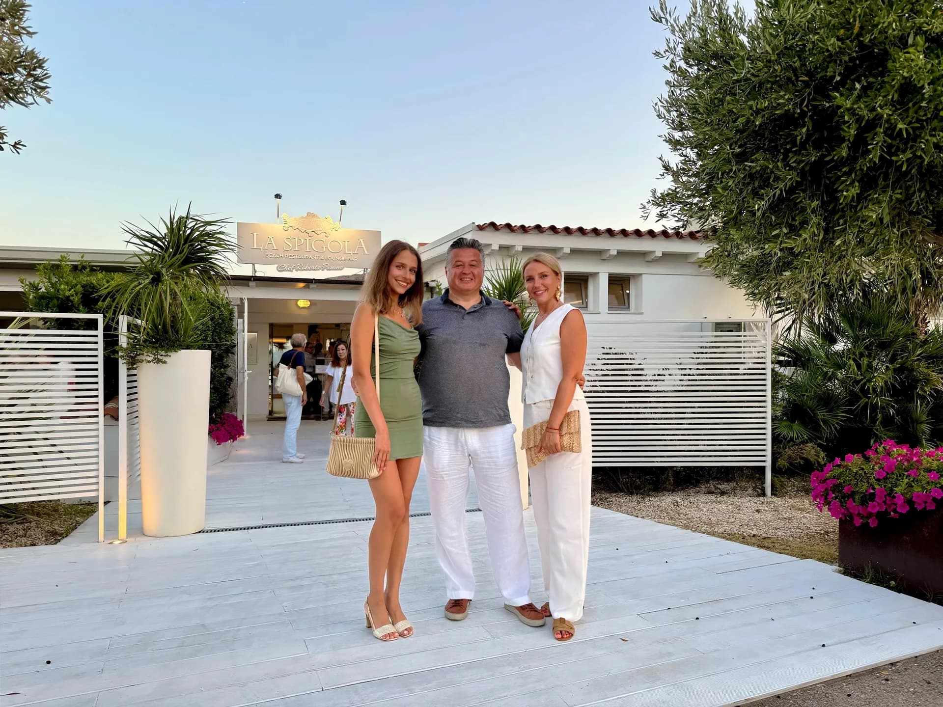 Tre persone sorridenti in abiti estivi in posa su una pedana di legno bianco davanti all'ingresso del ristorante in spiaggia La Spigola al crepuscolo, con ulivi e fiori rosa accanto a loro.